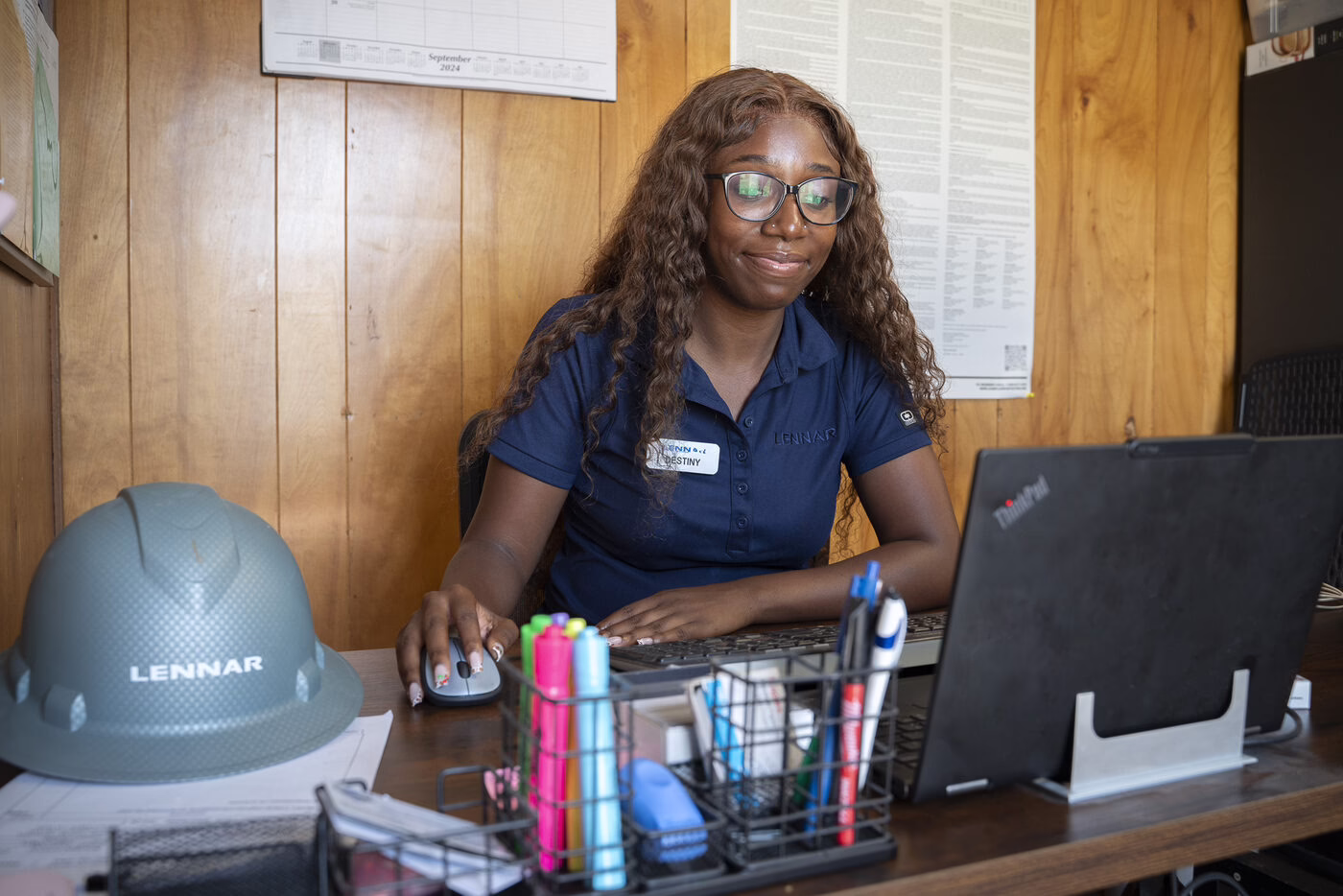 A young woman in a hard hat works at a desk at a housing organization.